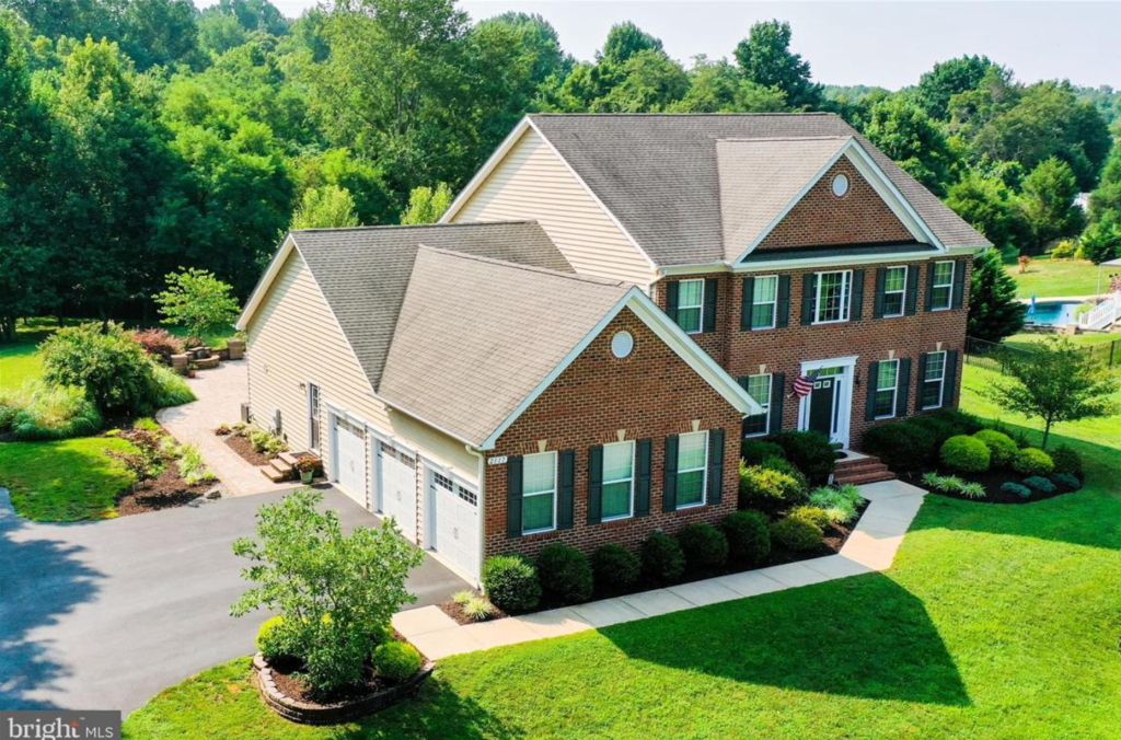 Aerial view of a large, modern brick house with a sloped roof, surrounded by green trees and landscaping. The property features a paved driveway, well-maintained garden, and a swimming pool in the background.