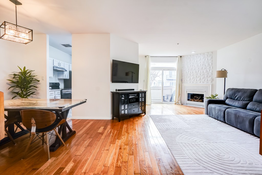 Modern living room and dining area featuring hardwood floors, a sectional sofa, a coffee table, and a flat-screen TV. Natural light enters through a large window with curtains.