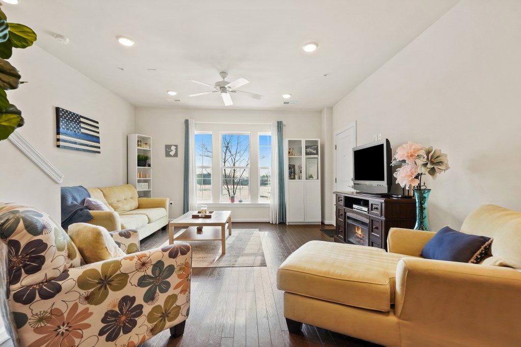 Cozy living room with large windows, a wooden coffee table, and a TV, featuring light-colored furniture and a decorative plant.