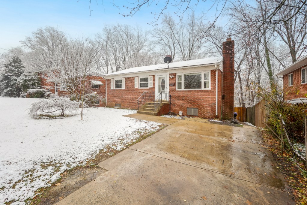 A brick ranch-style home with a driveway and steps leading to the front door, surrounded by a snowy yard and trees.