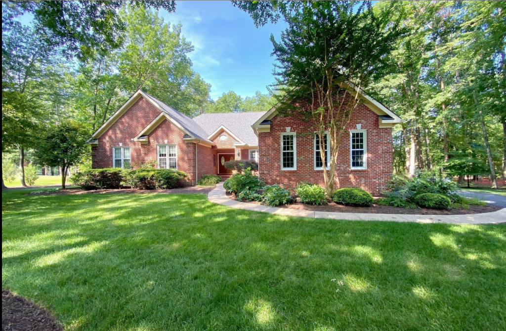 A brick house surrounded by lush greenery and trees, featuring a well-manicured front lawn and a curved pathway leading to the entrance.