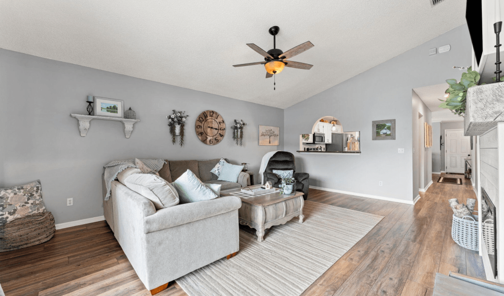 A cozy living room featuring a light gray color scheme, a ceiling fan, a comfortable sectional sofa, and decorative wall art, with a wooden coffee table in the center and a view of an adjacent entryway.