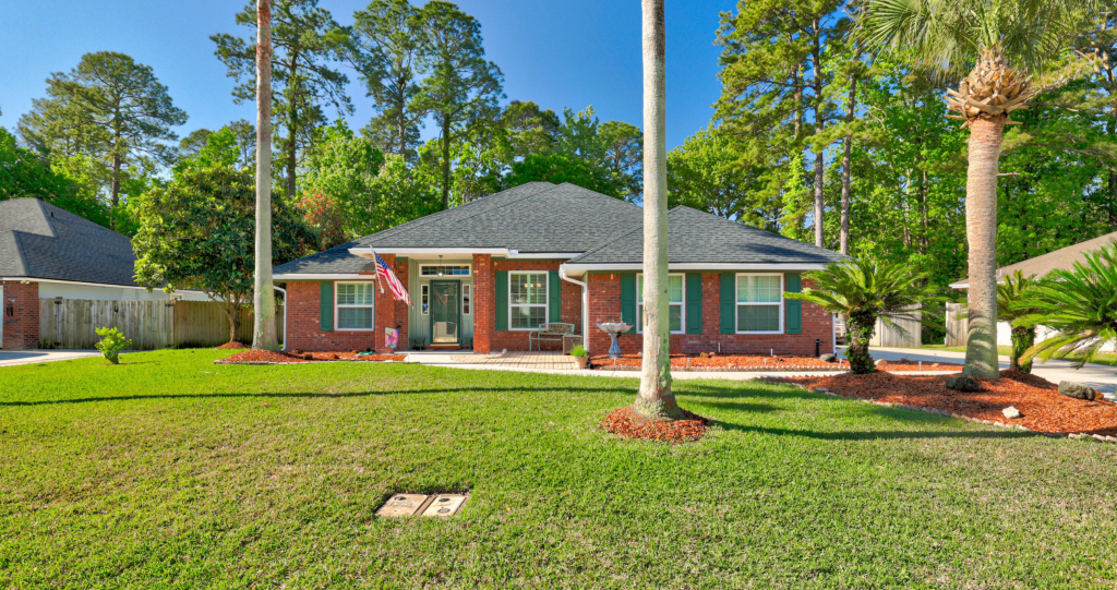 A charming brick house with a black roof, featuring a front porch, surrounded by a well-maintained lawn and palm trees. An American flag is displayed at the entrance, with a background of lush greenery.