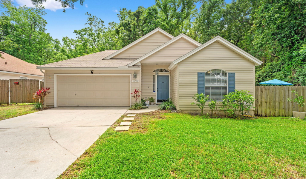 A single-story house with a beige exterior and a gray garage door, featuring a landscaped front yard with green grass and plants.