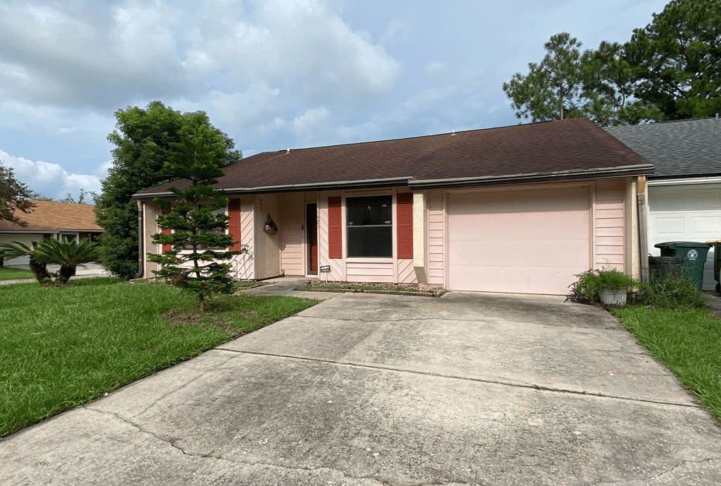 A single-story house with a pink exterior, featuring a brown roof and decorative shutters. The driveway is concrete, and there is a green lawn with trees on either side.