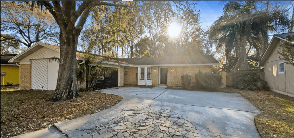 A sunny view of a single-story brick house with a driveway, surrounded by trees and greenery.